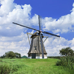 Ancient drainage windmill with thatched roofing in a green polder with dramatic shaped clouds and a blue sky.