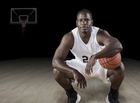 African American basketball Player holding a ball and posing in a darkened basketball arena.