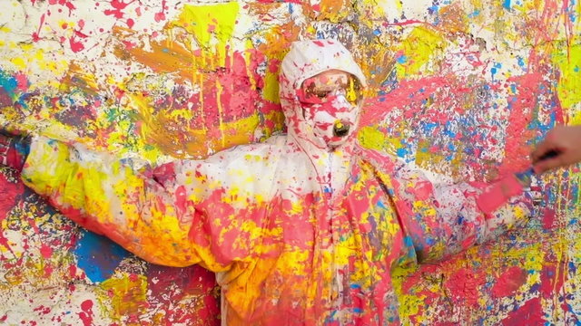 Person wearing protective coveralls standing by paint-splatter wall and being covered in multi-colored paint