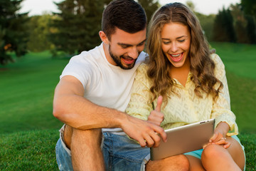 Guy and a girl sit on grass and talking on the tablet.