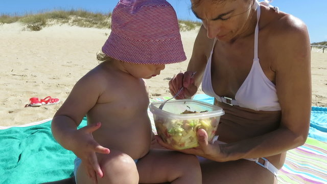 tender family two years age blonde baby with red white striped hat and blue swimwear with woman mother white bikini sharing eating pasta salad of tupperware sitting in beach