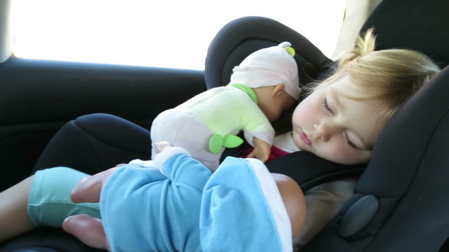 Tender Scene Of Blonde Caucasian Baby Face Two Years Old Age Sitting In Black Baby Car Seat Sleeping With Dolls In Her Arms, Vehicle Is Driving
