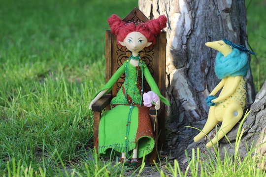Textile handmade doll in a green dress and burgundy hair sitting on a chair on a green glade