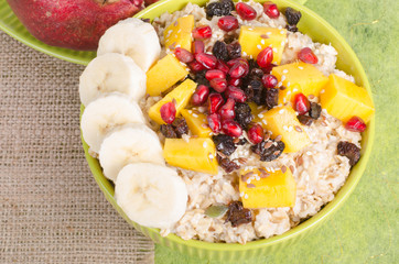 Oatmeal with mango, banana, seeds of pomegranate, raisins, dried fruits and sesame on canvas and green background. Healthy vegetarian breakfast.