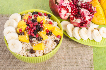 Oatmeal with mango, banana, seeds of pomegranate, raisins, dried fruits and sesame on canvas and green background. Healthy vegetarian breakfast.
