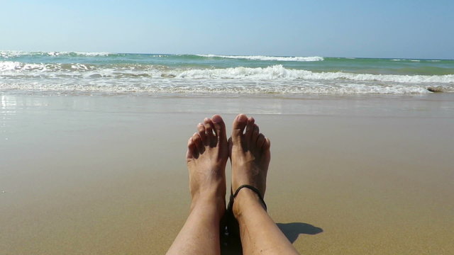 subjective shot of woman legs and bare feet sitting on golden sand beach looking at ocean water seaside and horizon in Cadiz Spain
