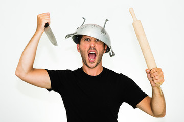 close-up of angry young caucasian man with a colander on his head and rolling pin and knife on his hands looking and screaming at you - isolated on white background - obrazy, fototapety, plakaty