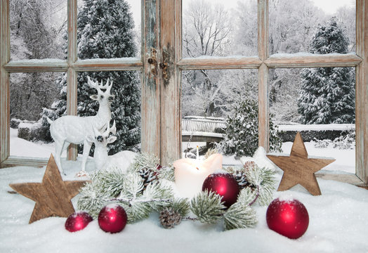 Stimmungsvolles Kerzenlicht in einem weihnachtlich dekorierten Winterfenster mit Blick in einen park&auml;hnlichen Garten mit Schnee