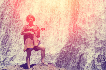 Young girl with ukulele, standing on a rock near waterfall