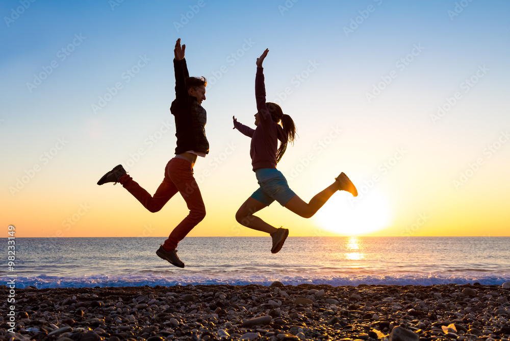 Wall mural Girl and Guy jumping high with arms up spectacular sunrise at ocean coast - Wall murals