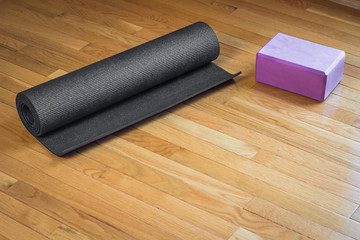 A black yoga mat rolled up next to a pink yoga brick on a wood floor