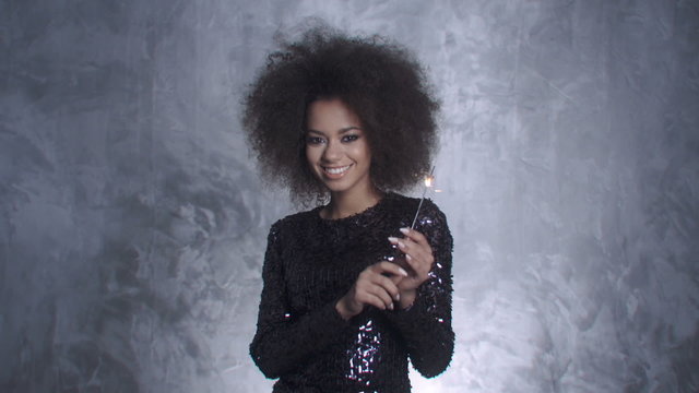Young African American woman posing in an elegant sparkling dress against silver background in a studio.