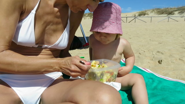tender family of two years age blonde baby with red and white striped hat with woman mother white bikini sharing and eating pasta salad of tupperware sitting at beach