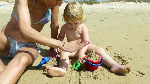 tender family two years age blonde baby green swimwear with woman mother bikini sitting in beach earth ground playing with sand red bucket and blue and green shovels trying to make castle