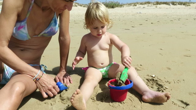 tender family two years age blonde baby with green swimwear with woman mother bikini sitting on beach earth ground playing with sand red bucket and blue and green shovels trying to make a castle