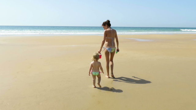 tender family scene of two years age blonde baby with green swimwear with brown hair woman mother blue bikini with red bucket in her hand walking on beach sand seaside with blue ocean and sky