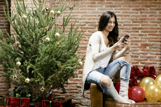 Brunette woman using her phone sitting by the Christmas tree at home.