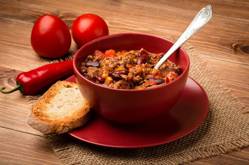 Chili con carne served in the red bowl on the wooden background.