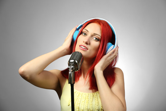 Beautiful young woman with microphone and headphones on gray background