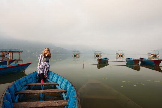 Happy meditate girl with blanket sitting in old boat on mountain lake early morning. Clouds and fog over the lake, loneliness, tranquility, relaxation
