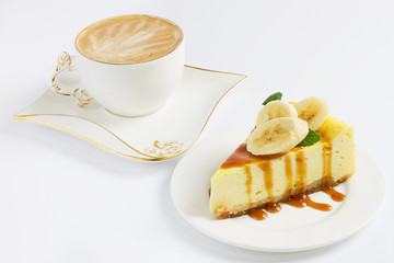 Slice of cheesecake with banana and caramel sauce, cup of coffee on the white background