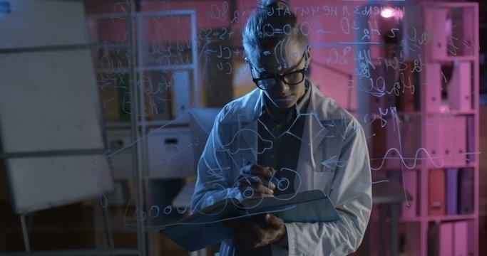 Young talented chemistry student reading book and writing molecular formulas on glass wall