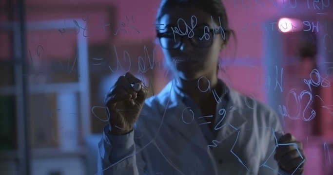 Young female scientist thinking about chemical formula and writing it on glass wall