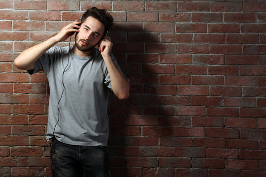 Young handsome man in grey T-shirt listening music with headphones on brick wall background
