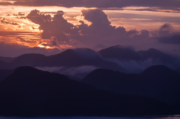 Obraz premium Silhouette of Mountains Above the Water under purple orange cloudscape clouds at sunset