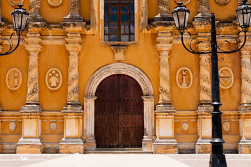 Old latern in front of yellow church with big brown wooden door, stone scarving and glass window