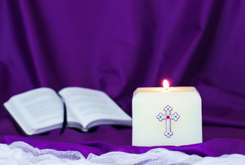 White Easter candle burning by bible and purple fabric blurred in background