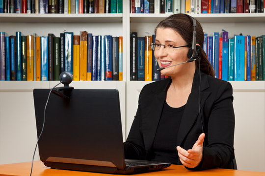 Teacher, tutor or professor with headset, laptop and camera in her office explaining something at an online lesson or video lecture, webinar.