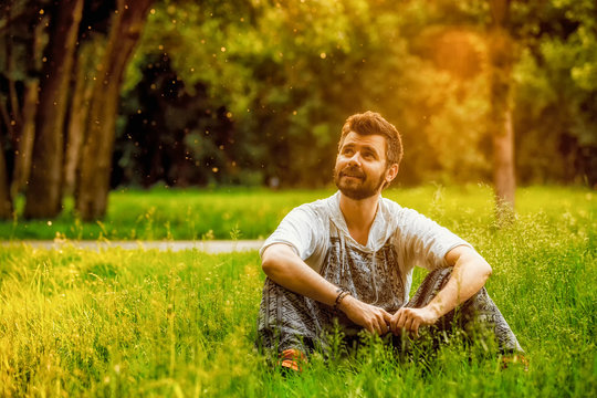 A bearded man sitting on a grass in the park on summer day, looks optimistic and relaxed