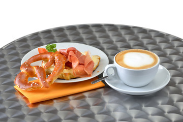 Snack bestehend aus Cappuccino und  Butterbrezel mit Parmaschinken, serviert auf Bistrotisch