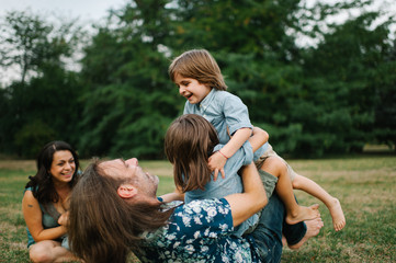 Happy young hipster family having fun while running, bowl,  rising up, piggyback ride their children in park on summer sunset
