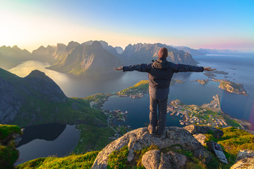 Silhouette of the man spreading arms and standing high on rock like the Statue of Christ the Redeemer looking at breathtaking view over small village,mountain landscape, sea during sunset in Norway