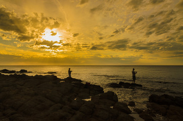 Silhouette two adult men fishing from rocks at sunset