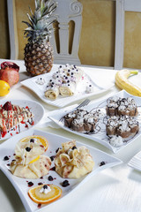 Four servings of pancakes with a variety of filling and decoration served on the table next to fresh fruit.