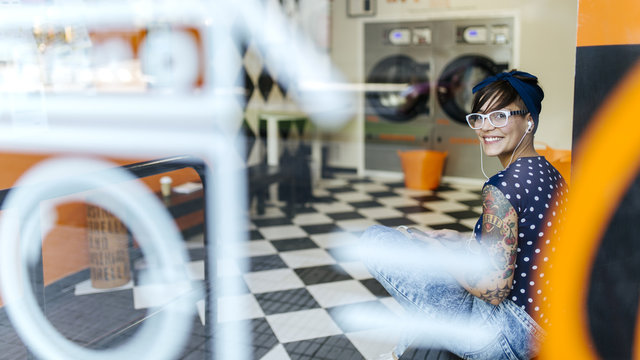 Portrait of tattooed young woman hearing music with earphones in a launderette