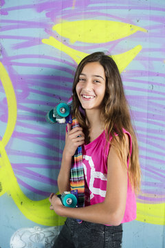 Portrait of smiling teenage girl holding a colorful skateboard in front of wall with graffiti