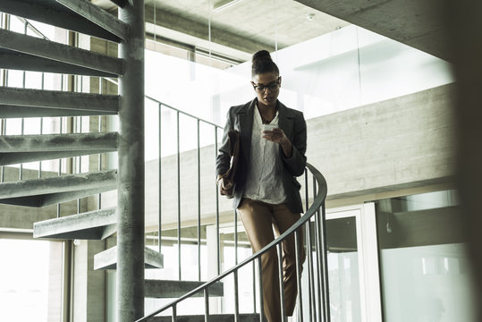 Young woman in office walking down spiral staircase looking on cell phone - Powered by Adobe