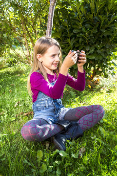 Little girl sitting on a meadow in the garden taking a selfie with smartphone