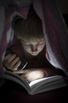 Boy with torch reading book under a blanket