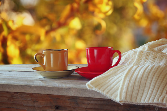 front image of two coffee cups over wooden table and woolen sweater in front of autumnal sunset background. Valentines Day concept
