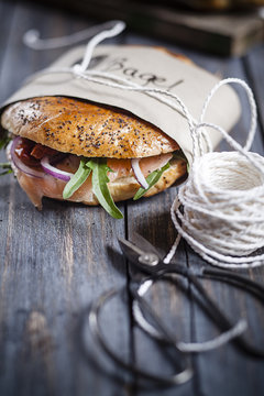 Two garnished home-baked bagels wrapped in paper, string and scissors on grey wood