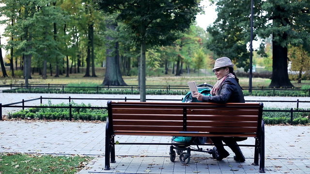 Happy mother with stroller chatting on tablet computer in the park
