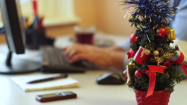 Small Christmas tree standing on the desk against a man typing on the computer 
