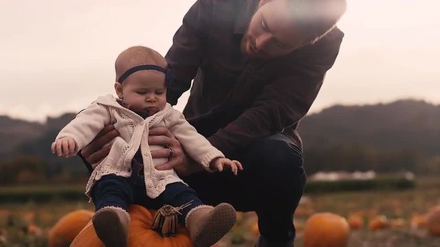 A baby sitting on a pumpkin at a pumpkin patch, with her dad holding her up