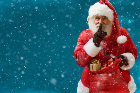 Santa Claus with huge red sack keeping forefinger by his mouth and looking at camera / Merry Christmas & New Year's Eve concept / Closeup on blurred blue background.