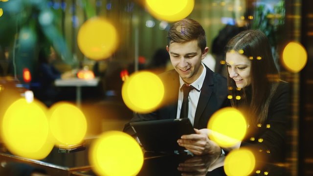 Young Couple Sitting In A Cafe And Looks At The Screen Of The Tablet